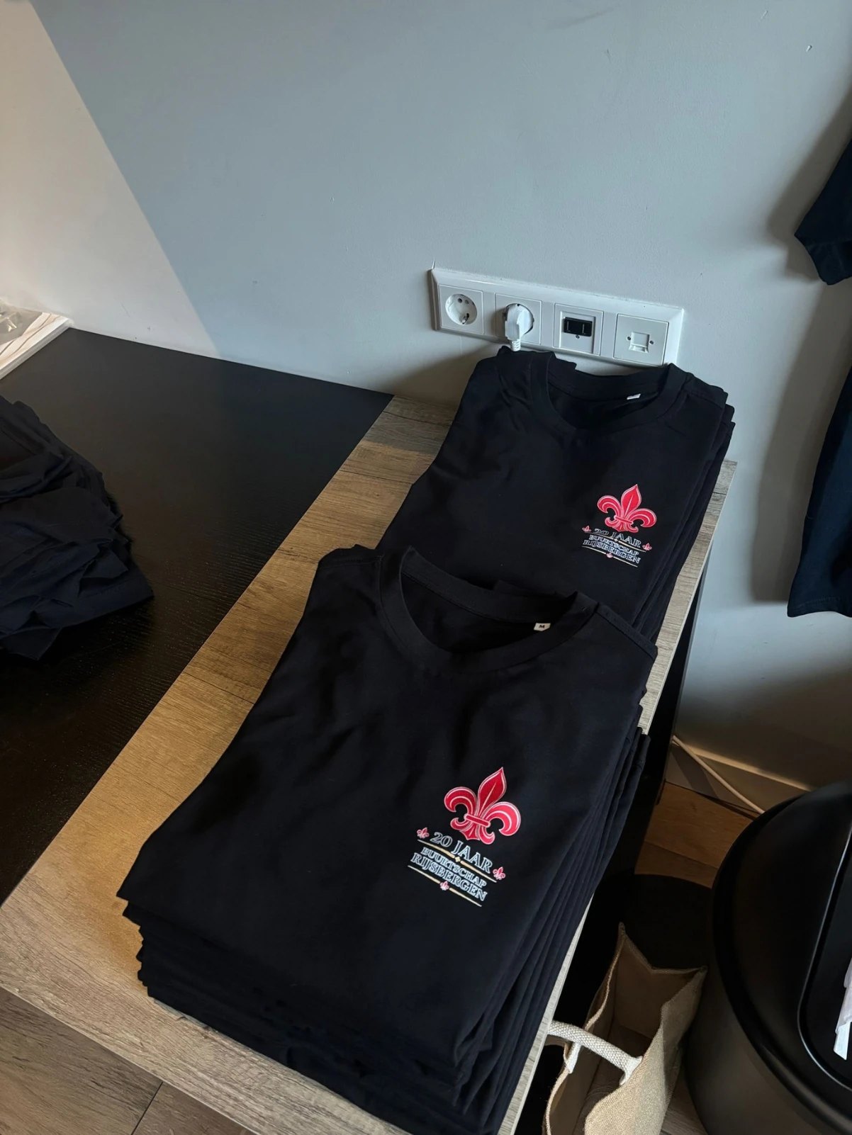 Stapels zwarte shirts met een rode Franse lelie en de tekst 20 jaar Buurtschap Rijsbergen